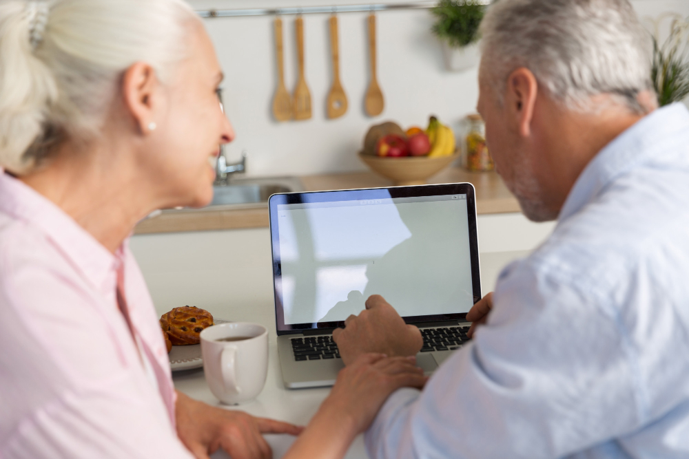 Twee ouderen mensen zitten aan de keukentafel achter een laptop.