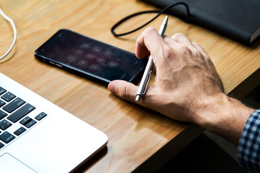 Een persoon heeft een pen in zijn hand met naast hem een laptop en een mobiel.