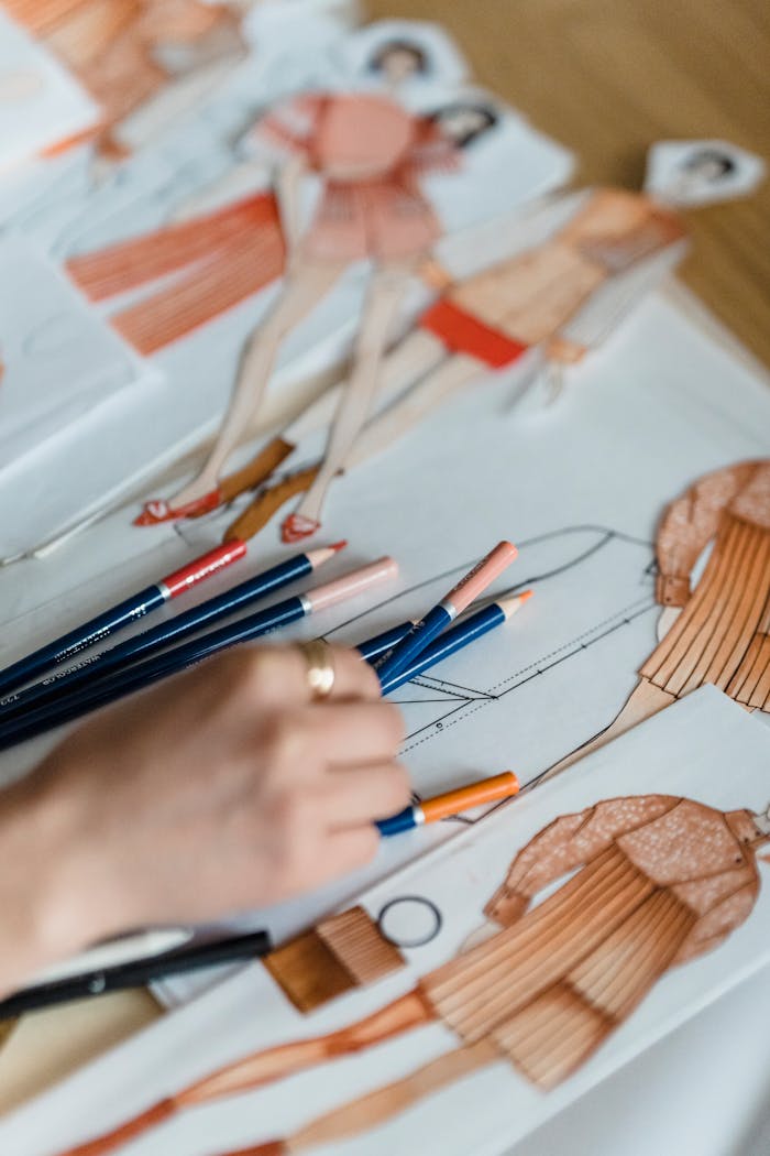 High angle of crop anonymous illustrator working at desk with colored pencils and cutout human figures in outfit