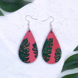 Pair of hand painted earrings with coral background and green monstera leaves in the foreground. By DSB Designed.