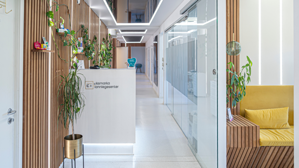 A modern and welcoming reception area at Eiksmarka Tannlegesenter featuring wooden panel walls adorned with green plants, a sleek white reception desk, and a cozy yellow bench enhancing the calm and inviting atmosphere for visitors.