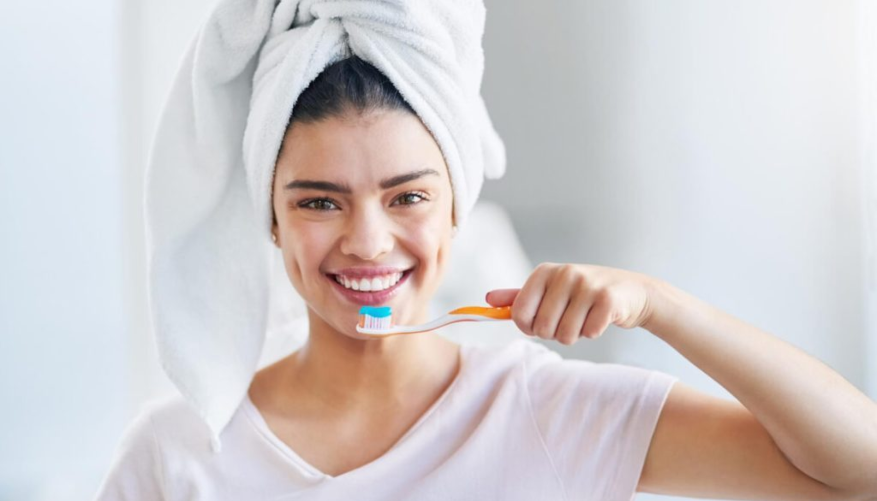 Smilende kvinne med håndkle på hodet holder en tannbørste med tannkrem, klar til å pusse tennene på badet.