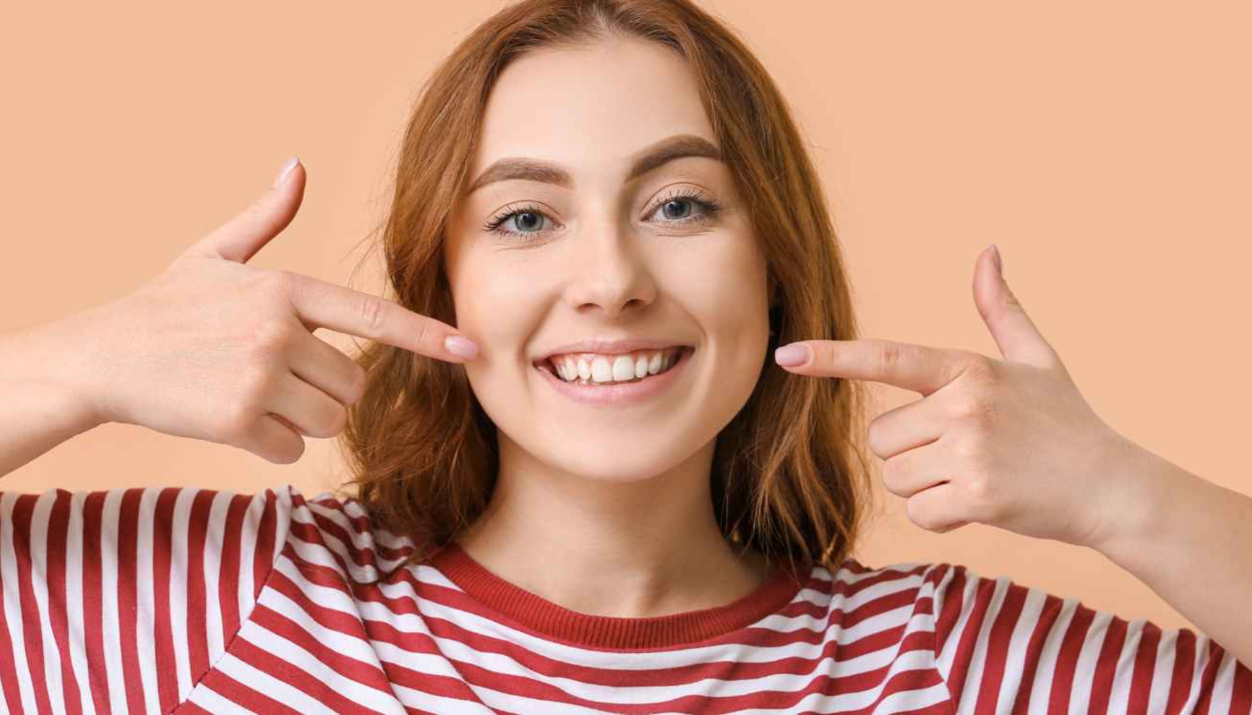 En kvinne smiler og peker på tennene sine med begge hender, som viser et vakkert smil.