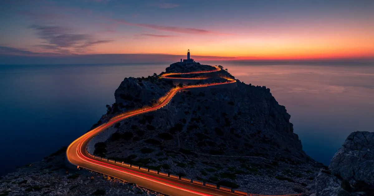 Winding coastal road illuminated at sunset leading to Formentor Lighthouse on rocky cliff, overlooking Mediterranean Sea in Mallorca.