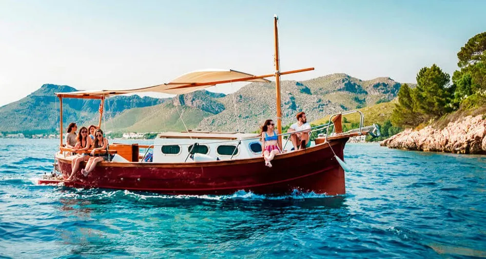 Group of friends enjoying a sunny boat trip on a traditional wooden sailboat in the Mallorcan side of Mediterranean Sea, with rocky mountains in the background