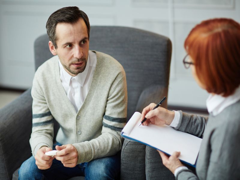 Depressed middle-aged patient sitting nervously on comfortable armchair with handkerchief in hands and explaining his problems to mature psychologist with clipboard and pen
