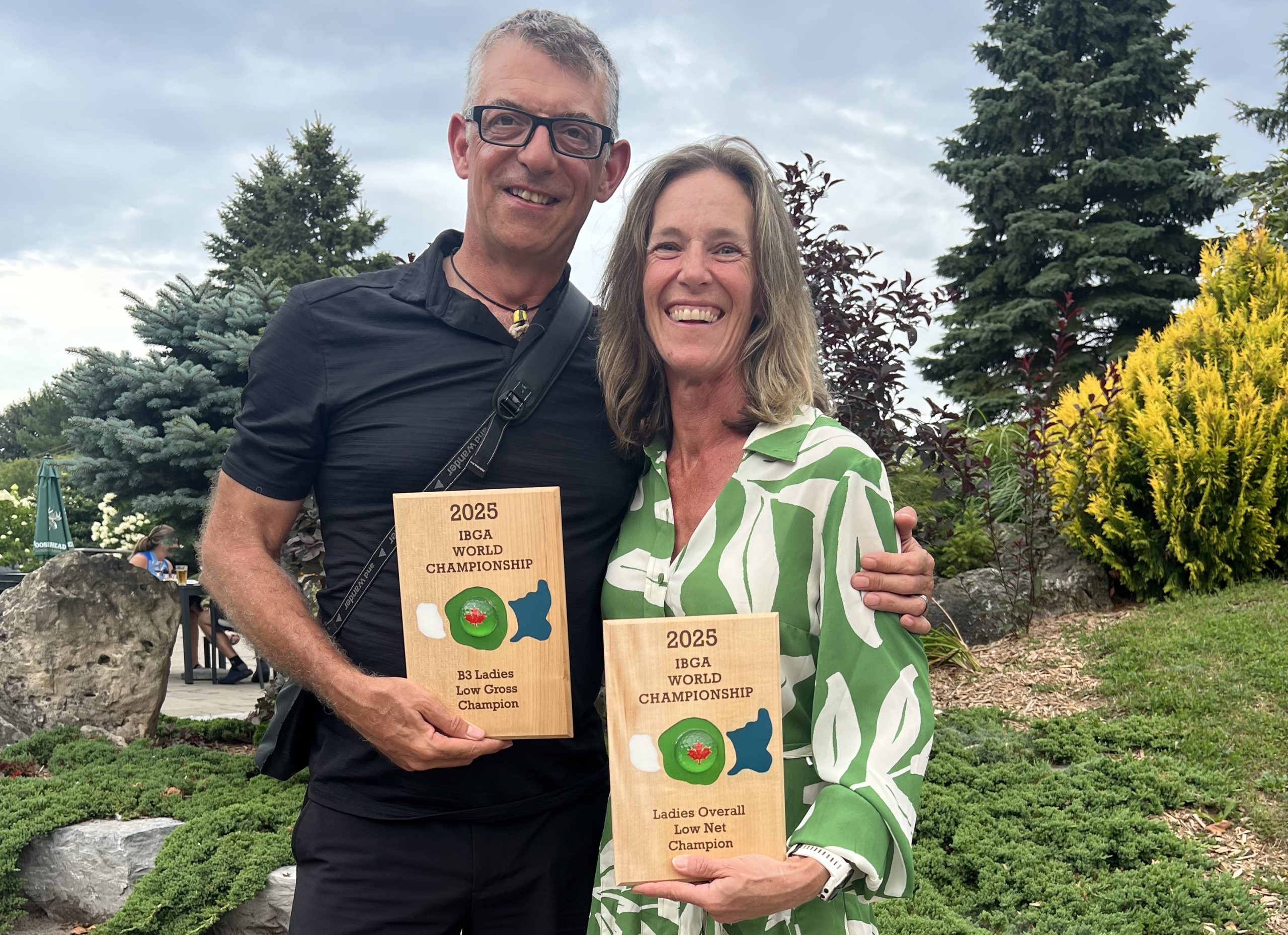 Photo shows Mandy in a delightful green and white dress with husband John, holding Wooden plaques with their awards engraved.