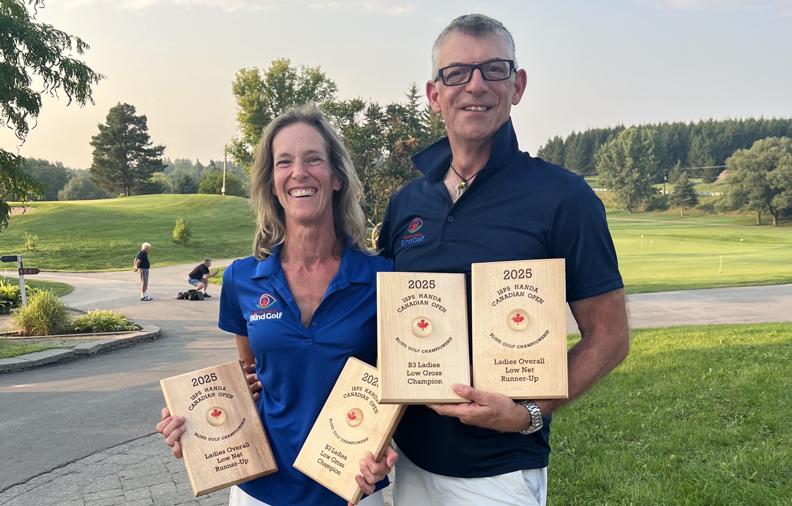 Photo shows Mandy and John holding wooden plaques inscribed with the Canada Open and the ladies B2 and overall prizes.