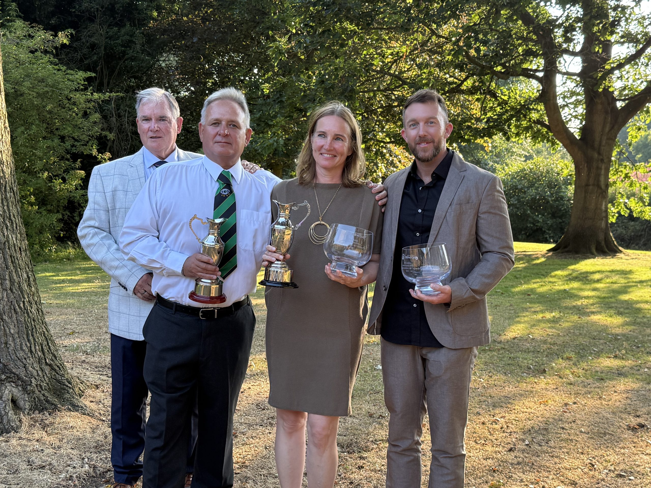 Photo shows Karin and and Owen with their claret jugs. The are accompanies by their guides and are in a grassy area with trees behinde