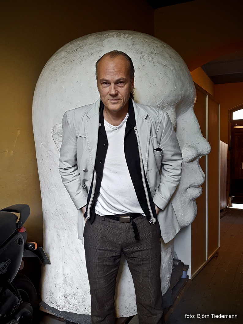 BILD: Håkan Sandell stående i ljus kavaj framför ett stort huvud, foto: Björn Tiedemann