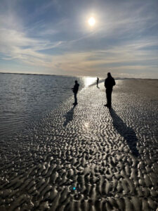 3 personer står på en strand. Ansigter er ikke genkendelig, solen skinner i det fjerne og laver skygger på strand sandet.