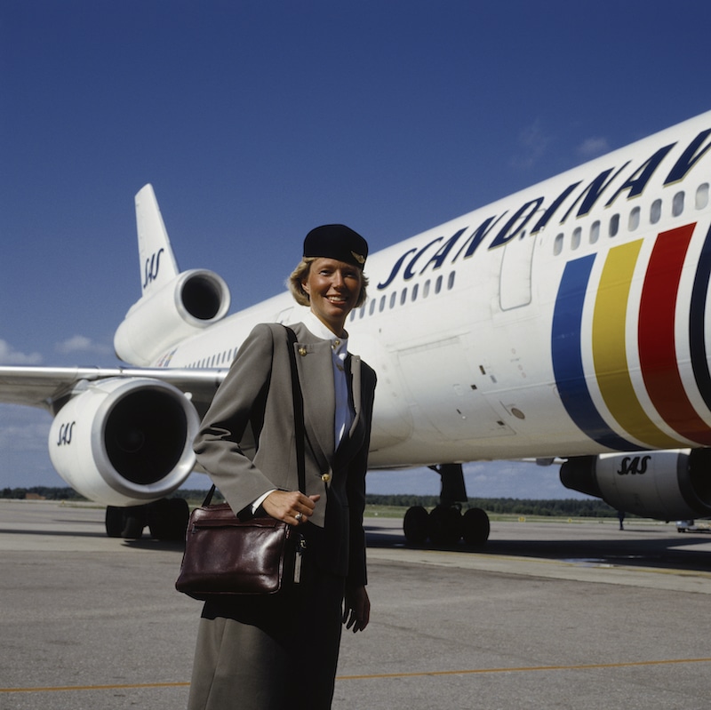 SAS Uniforms. Air hostess in summer uniform with cap and shoulder bag in bordeuax coloerd leather designed by Calvin Klein 1983-1999. Air hostess on the front of SAS aircraft.
