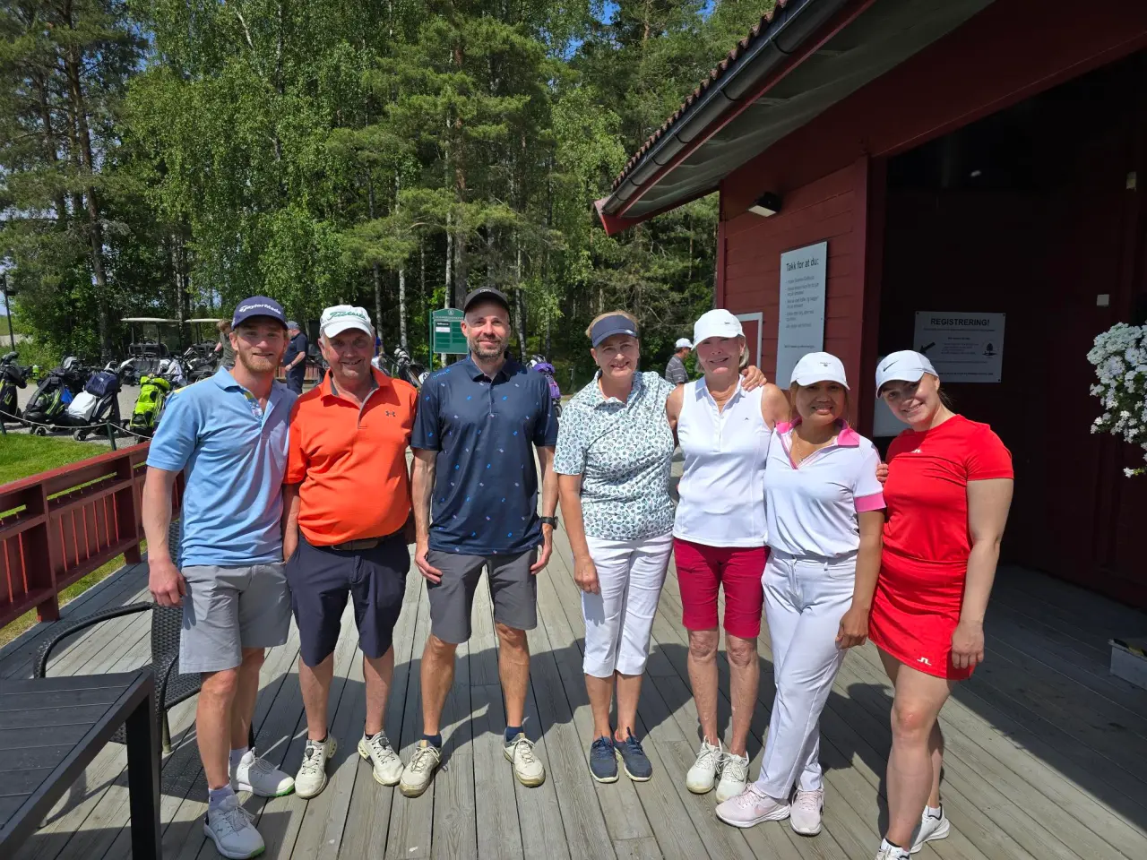 Tre herrer og fire kvinner står i golftøy på klubbhusets terrasse og smiler til kamera i solen.