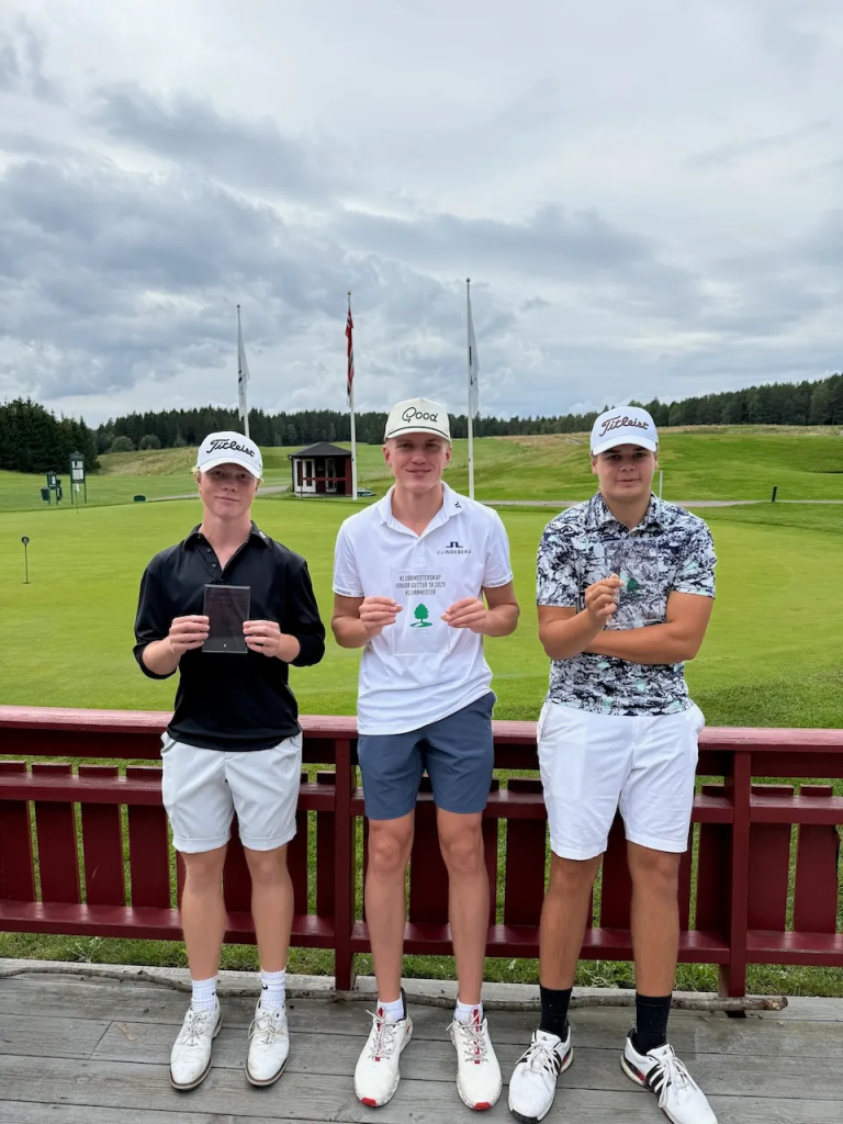 Tre unge menn i shorts, caps og skjorter står på klubbhusets terrasse og ser i kamera og smiler mens de holder en firkantet glasspokal. Bak dem ser vi Grønmo golfbane. 