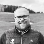 Mann med veldig kortklipt hår, briller med firkantede, sorte rammer og skjeggvekst smiler og ser i kamera.