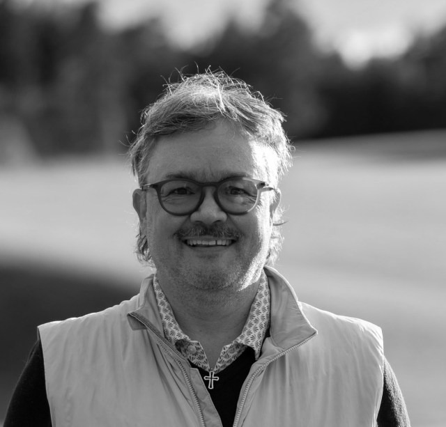 Mann med kort hår og briller med sort ramme, lys vest og skjorte smiler og ser i kamera. 