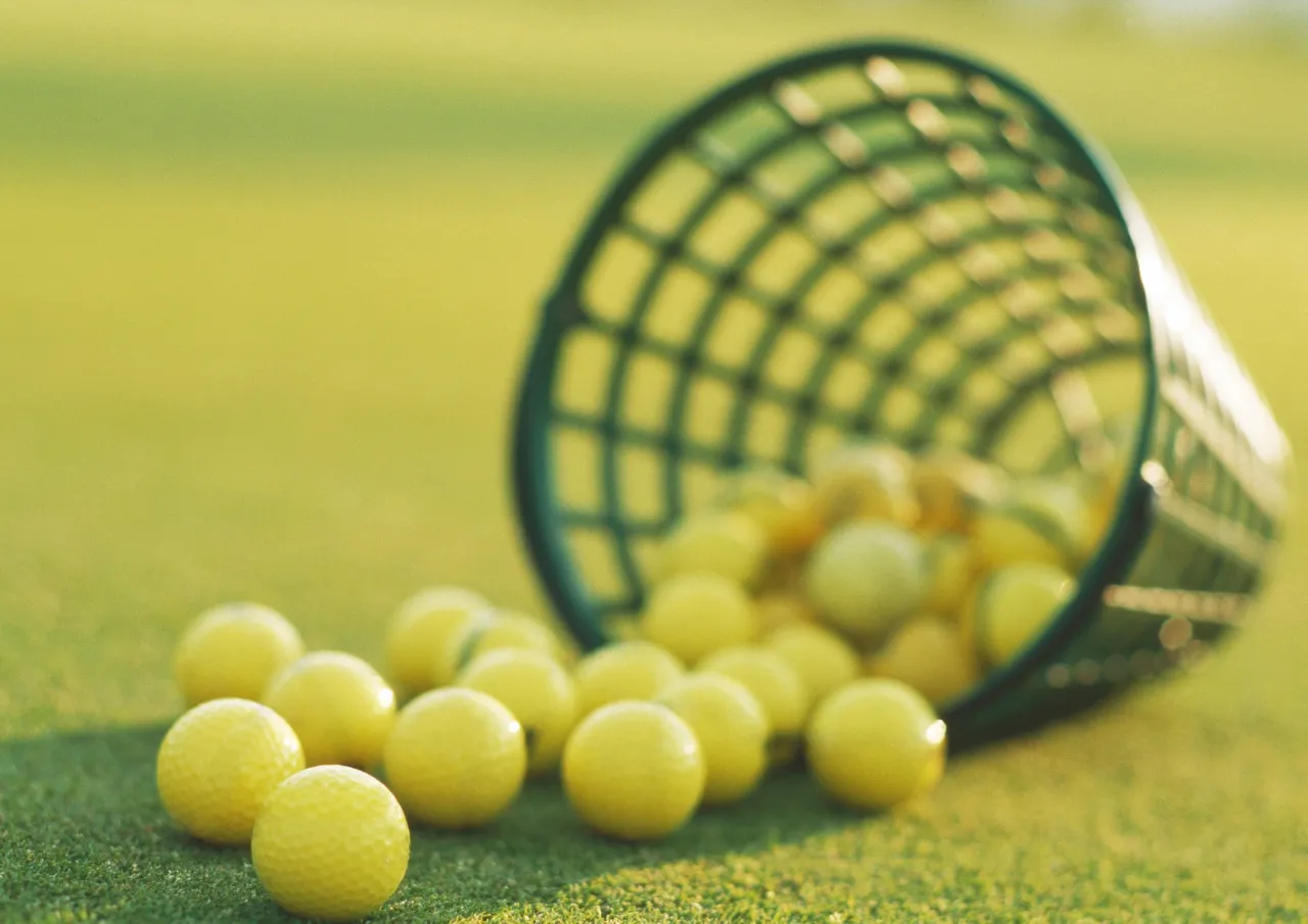 En grønn kurv ligger veltet, og en rekke gule golfballer er både på innsiden og utsiden av kurven på en kortklipt, grønn plen.