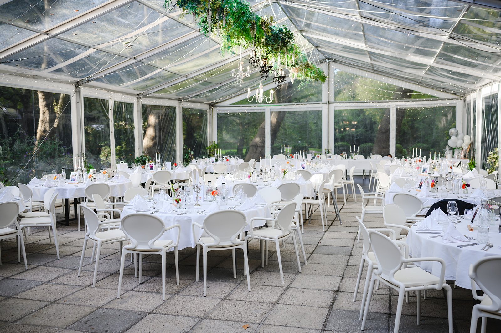 Lichtdurchflutetes Zelt mit weiß gedeckten Hochzeitstischen, Hängegrün und runder Bestuhlung – elegante Outdoor-Hochzeitslocation im Grünen.