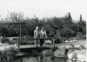 Foto af Jutta og Peter Ottesen stående ved rækværket på en bro over vandløbet fra en lille sø i deres smukke have.