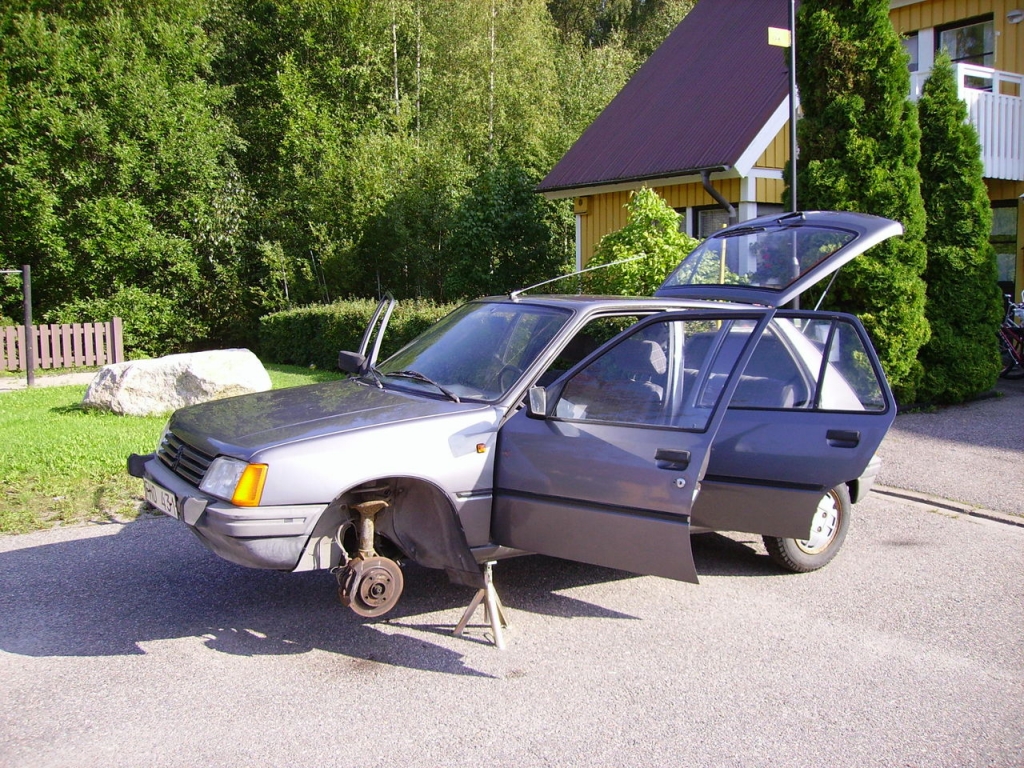 Hej Bruksbils presentatör Theo's första bil, en antracitgrå Peugeot 205 GR från 1985. Står på pallbockar framför ett gult hus.