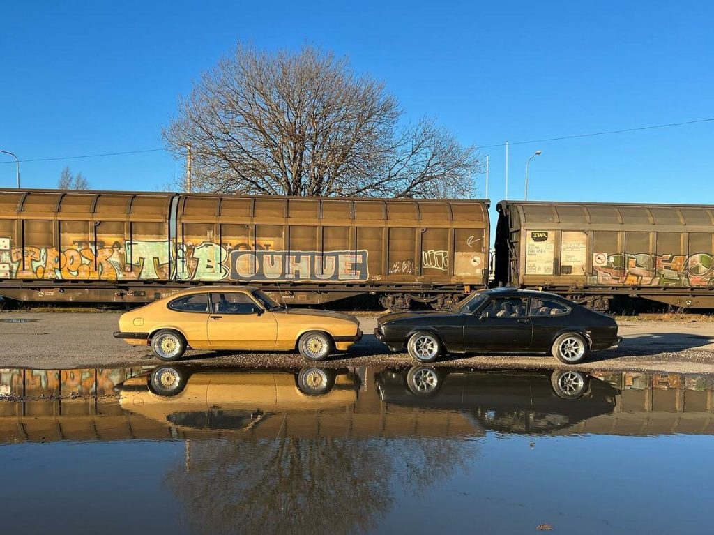 Två Ford Capri MK3 vid en vattenpöl, framför tågvagnar.