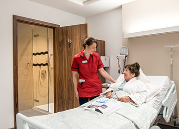This photo shows a professional carer tending a patient that lies in bed in a private room. The patient is comfortable and has been reading a magazine. In the background is a private en suite with accessible shower.