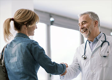 This photo represents a female ventral hernia patient meeting a surgical-coat-wearing and welcoming consultant surgeon. They are shaking hands.
