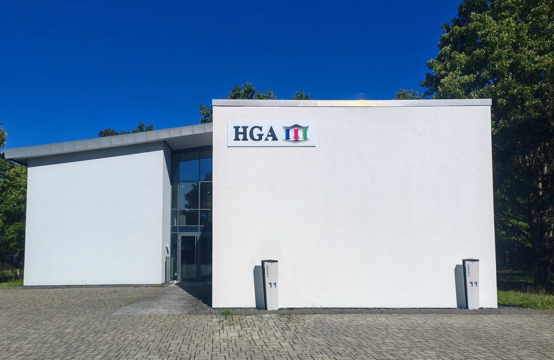 Außenansicht des weißen, zweistöckigen HGA-Hessen-Gebäudes in der Fritz-Remy-Straße 25 mit Parkplatz, Bäume unter blauem Himmel.