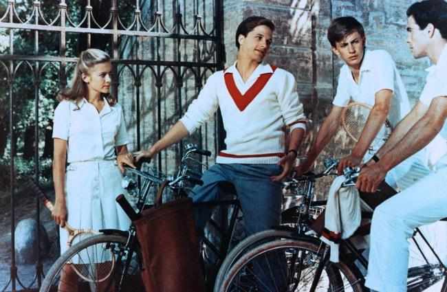 Tennisschläger in der rechten Hand steht vor einem Gittertor und hört drei Jungen Männern zu, die  rechts von ihr auf Fahrrädern sitzen und sich unterhalten.