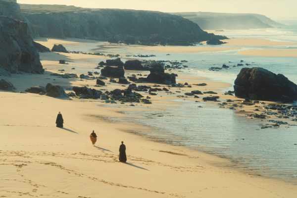 Drei dunkel gekleidete Menschen gehen in großem Abstand von rechts nach links über einen Sandstrand eine Felsküste am Meer entlang.