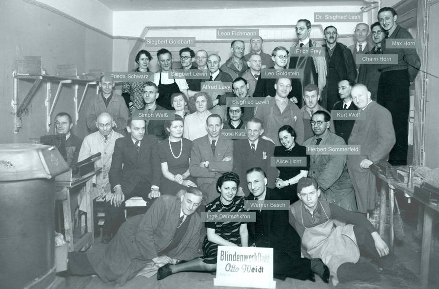 Das Schwarz-weiß-Foto zeigt 35 Männer und Frauen in einer Werkstatt in vier Reihen auf einer Tribüne aufgereiht. Einige von ihnen sind mit einem Namen versehen, vorne in der Mitte Inge Deutschkron.
