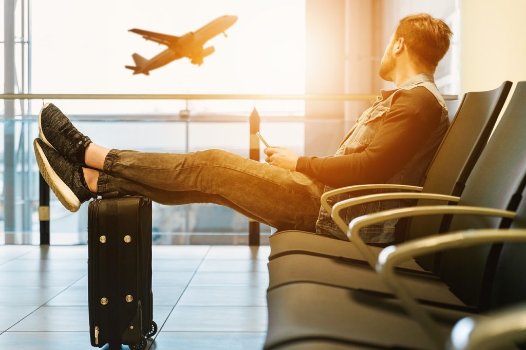Person som sitter avslappet på en flyplass med føttene på en koffert, mens et fly tar av i bakgrunnen under sollys.