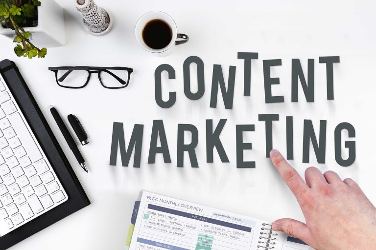 Et skrivebord med kaffekopp, briller, tastatur, penner og en notatbok, der en finger peker på teksten 'Content Marketing' – symboliserer strategisk innholdsproduksjon.