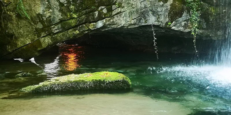 Vann som glitrer under fjellet ved Aggitis-grotten, med mosekledd stein og et lite fossefall som gir liv til landskapet.