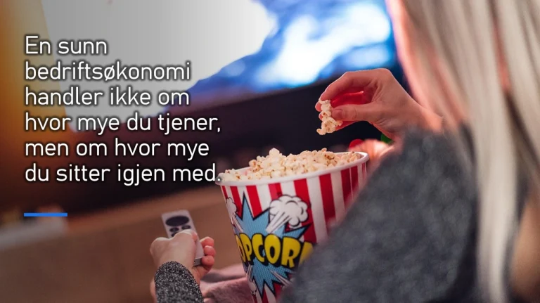 En person med langt, lyst hår sitter i en sofa og ser på TV mens de spiser popcorn fra en stor, rød-hvit bøtte med ordet "POPCORN" og en tegneserieaktig eksplosjon på. Personen holder også en fjernkontroll i den andre hånden. På venstre side av bildet er det en tekst i hvitt som sier: "En sunn bedriftsøkonomi handler ikke om hvor mye du tjener, men om hvor mye du sitter igjen med." Bakgrunnen er uskarp, og TV-skjermen lyser opp rommet.
