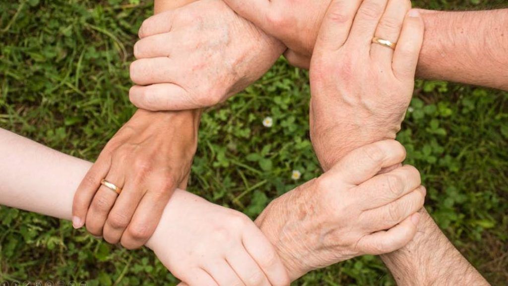 Seks hender i ulike hudtoner holder hverandre i et lukket kjedemønster over grønt gress, som symbol på samarbeid, fellesskap og samhold på tvers av ulikheter. Flere av hendene bærer gifteringer.