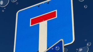 Nærbilde av et blått trafikkskilt som viser «blindvei» (T-symbol med rød ende), mot en mørk blå bakgrunn med vanndråper, muligens som en visuell effekt.