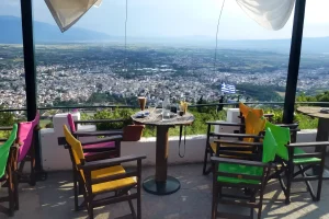 Utsikt fra en kaféterrasse over byen Drama i Nord-Hellas, med fargerike stoler, bord med drikke og det greske flagget i bakgrunnen. Foto: Bearbeidet fra Wikimedia Commons.