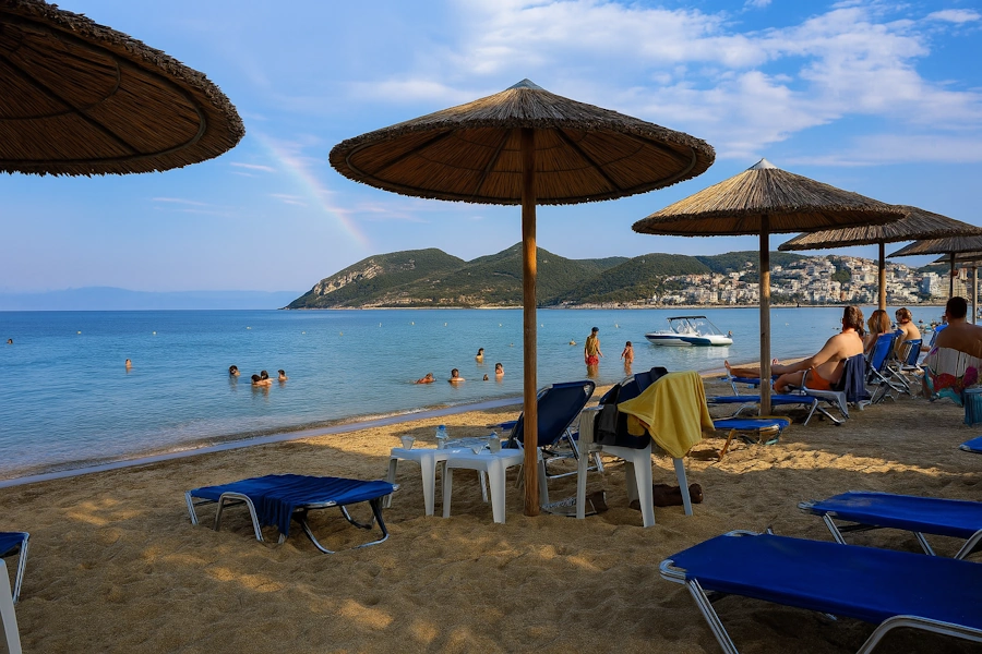 Strand med parasoller og solsenger i forgrunnen, folk som bader i det rolige havet, en liten båt i vannet og en åsside med hus i bakgrunnen. En svak regnbue skimtes over fjellene under en delvis skyet himmel. Foto: Raymond Sebergsen