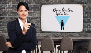 Confident businesswoman standing in a meeting room, holding glasses in hand, with a sign in the background that reads "Be a leader. Not a boss."