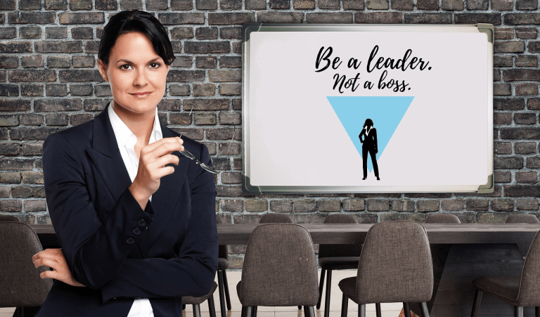 Confident businesswoman standing in a meeting room, holding glasses in hand, with a sign in the background that reads "Be a leader. Not a boss."