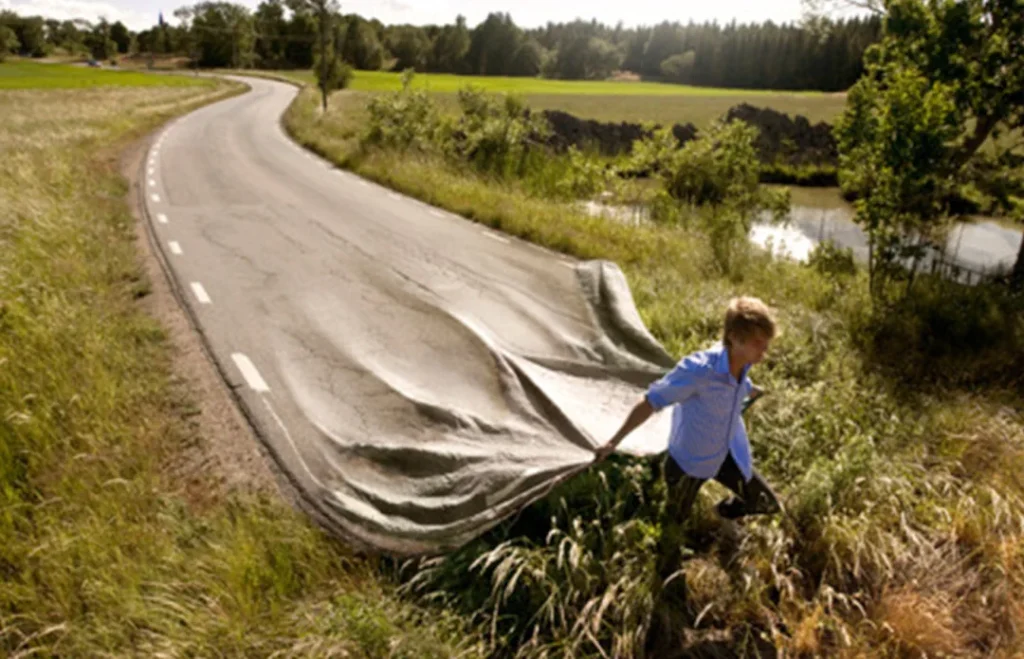 A person lifting the corner of a country road as if it were a carpet, revealing a new path beneath. Surrounded by fields, trees, and water in a rural landscape, the image symbolizes the idea that the path is created as we walk it, representing new perspectives, opportunities, and personal growth.