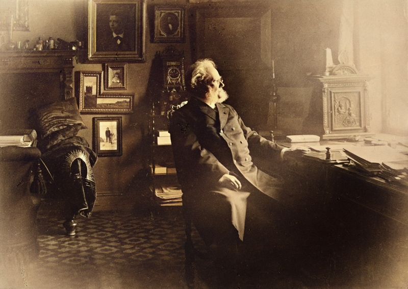 Henrik Ibsen sittende ved skrivebordet i leiligheten sin i Oslo, med lys som faller inn fra vinduet.