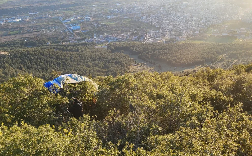 Paraglider som har landet i kratt og buskas under startområdet på Korylovos, med utsikt over byen og slettene nedenfor i ettermiddagssol.