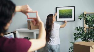 En person henger opp et innrammet bilde på veggen mens en annen måler avstanden med hendene. Flytteesker og planter står i rommet.