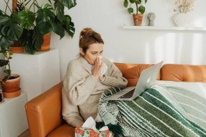 Sick woman working remotely from home on a couch, wrapped in a blanket, using a laptop while blowing her nose, surrounded by plants and tissues.