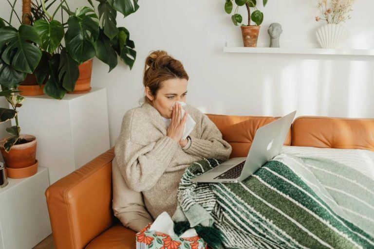 Sick woman working remotely from home on a couch, wrapped in a blanket, using a laptop while blowing her nose, surrounded by plants and tissues.