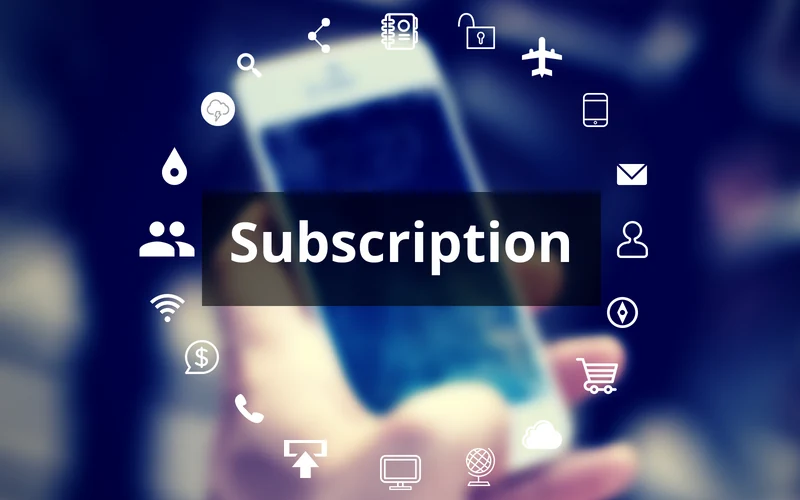 En uskarp bakgrunn med en hånd som holder en mobiltelefon. I forgrunnen står ordet 'Subscription' i hvit skrift på en svart rektangulær boks. Rundt teksten er en sirkel av hvite ikoner som representerer ulike digitale tjenester, som skytjenester, e-handel, e-post, kommunikasjon og nettverk, noe som symboliserer abonnementstjenester i en digital kontekst.