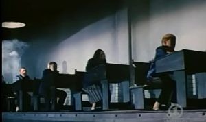 En scene fra filmen Pink Floyd – The Wall som viser skoleelever sittende ved identiske pulter i et dystopisk, fabrikk-lignende klasserom. Lyset og skyggeleggingen skaper en kald og undertrykkende atmosfære, som understreker filmens kritikk av et rigid utdanningssystem der elevene behandles som produkter på et samlebånd.