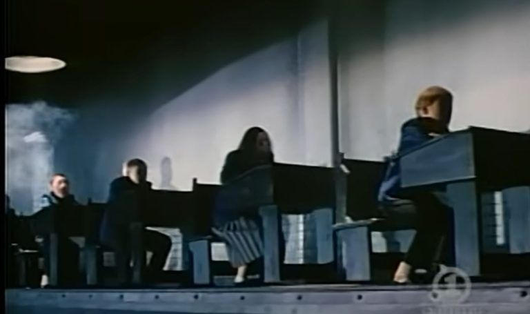 En scene fra filmen Pink Floyd – The Wall som viser skoleelever sittende ved identiske pulter i et dystopisk, fabrikk-lignende klasserom. Lyset og skyggeleggingen skaper en kald og undertrykkende atmosfære, som understreker filmens kritikk av et rigid utdanningssystem der elevene behandles som produkter på et samlebånd.